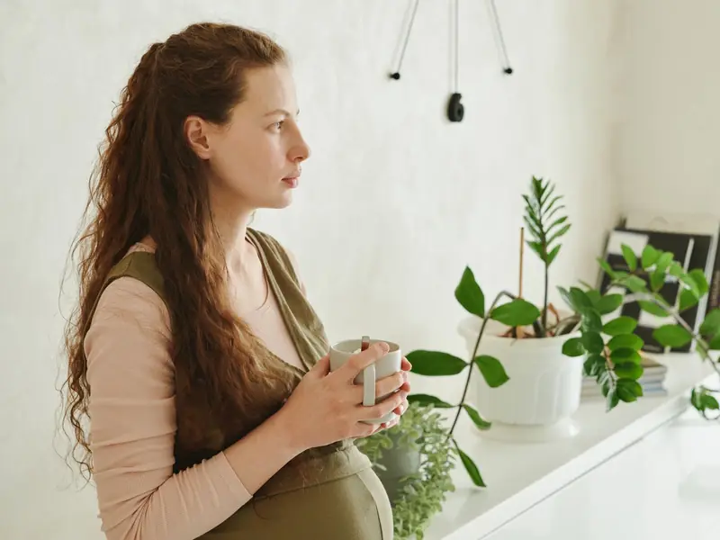 A pregnant woman holds a mug of tea and looks out of a window in a bright room with houseplants.